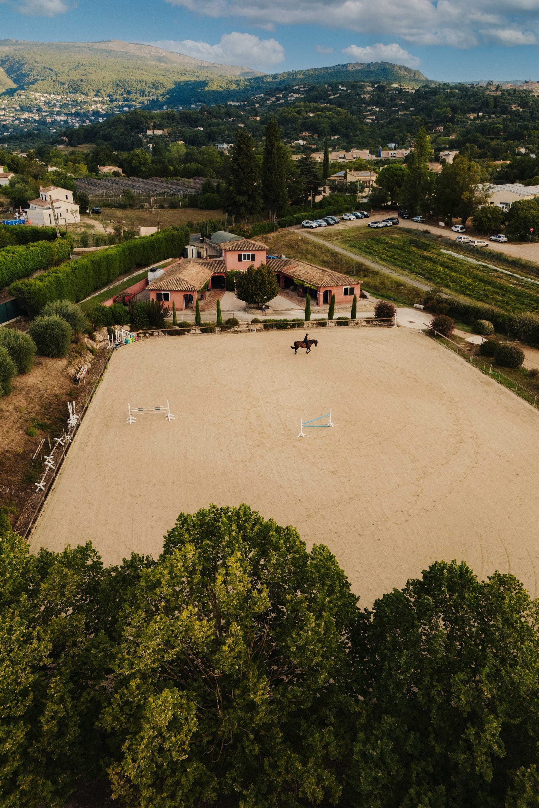 Aerial view of the pink stables and sand riding arena among the trees, with the hills of Grasse behind